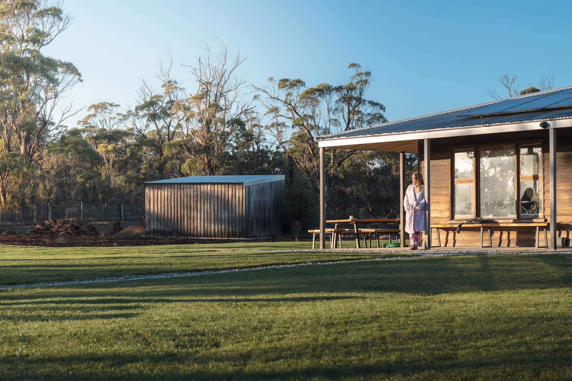 A woman in a dressing gown stands on the veranda of a wooden house, with a shed and trees in the background and sunlight casting shadows on the grass.