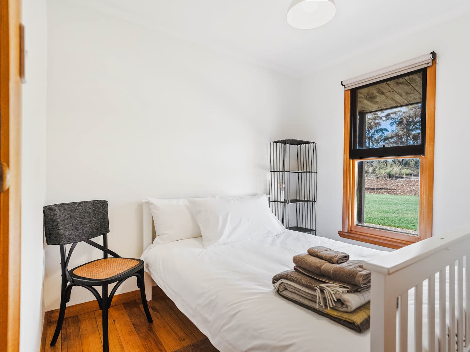 A simple bedroom with a white double bed, stacked towels, a black chair, a cot, wooden floors, and a window showing greenery outside.