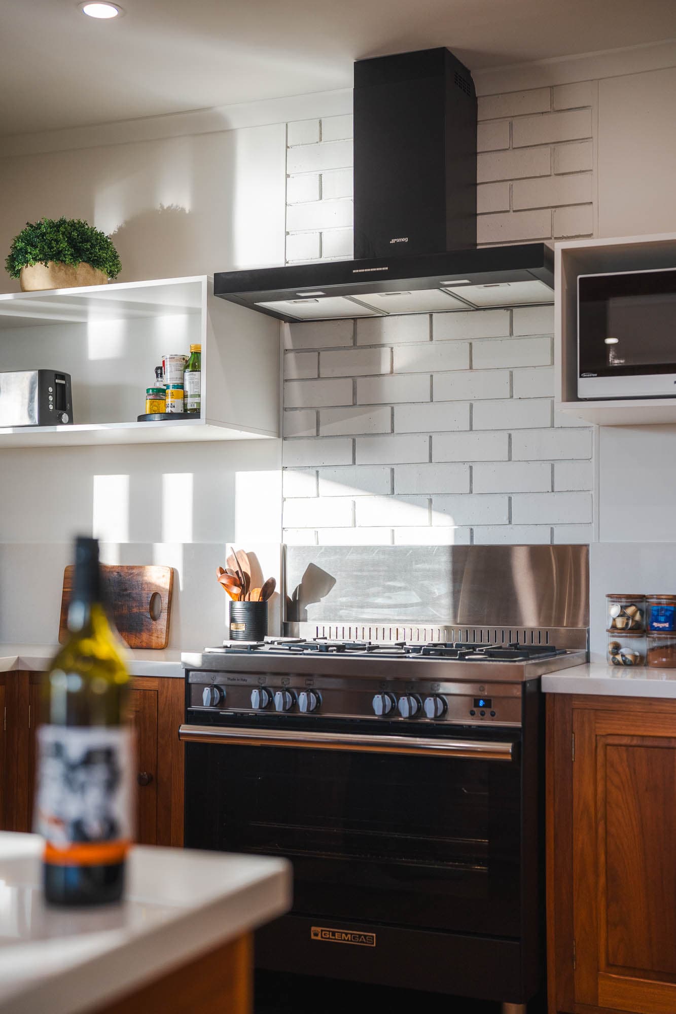 Modern kitchen with a black cooker hood, gas hob, white brick splashback, wooden cupboards, open shelves, and a wine bottle on the worktop in the foreground.