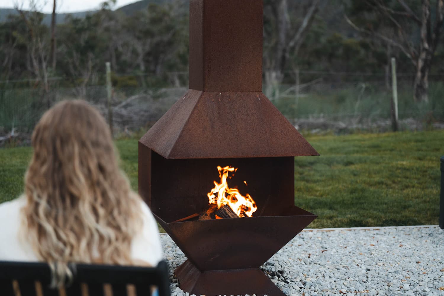 A person with long wavy hair sits on a chair facing an outdoor metal fire pit with a burning fire, in a garden with grass and trees.