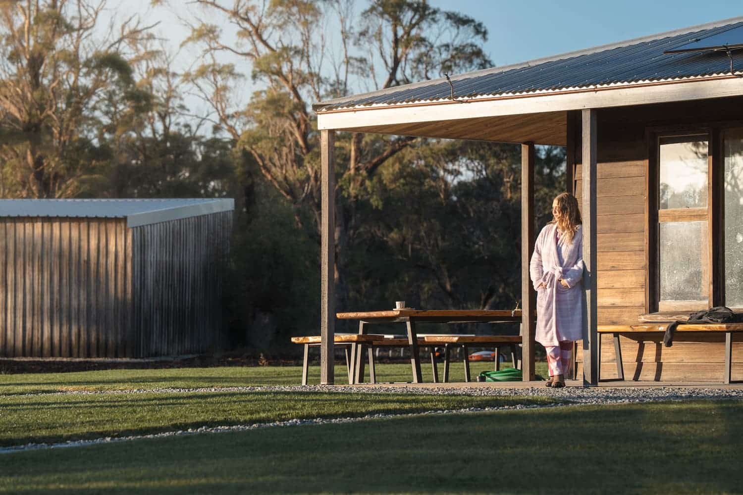 A person in a dressing gown stands on the veranda of a wooden house, looking out towards the garden in early morning light.