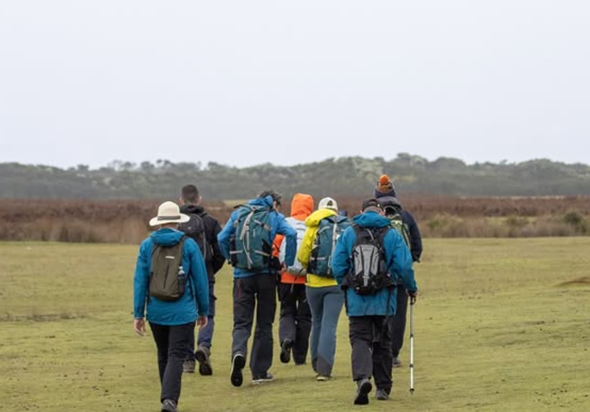 A group of seven walkers wearing outdoor gear and rucksacks walk across a grassy field under an overcast sky.