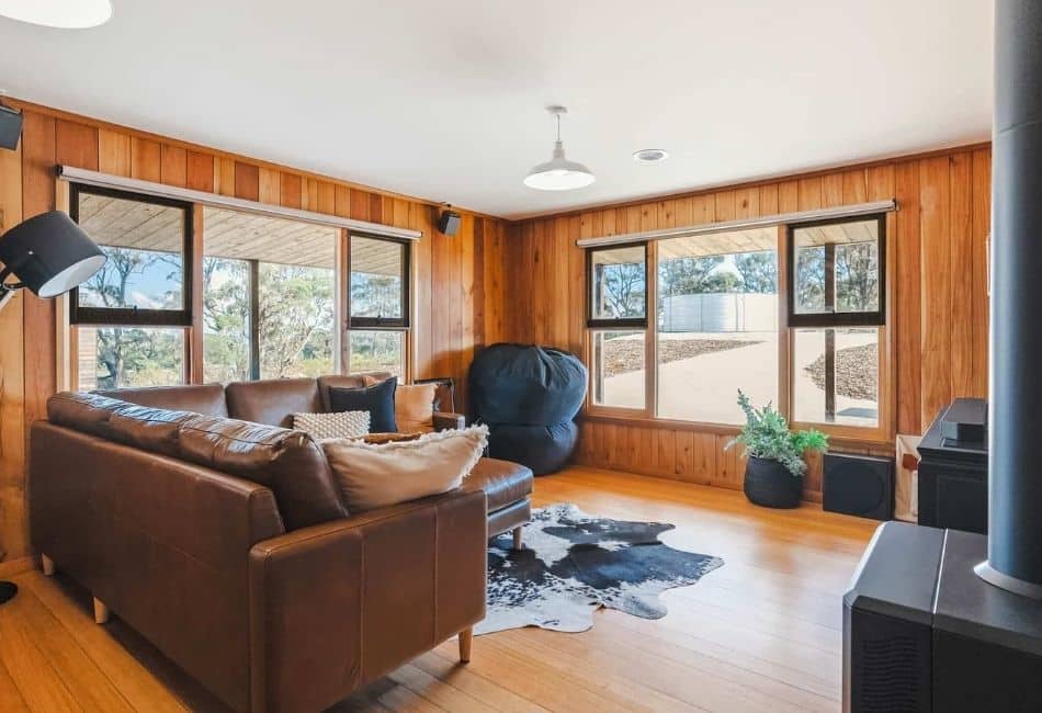 Living room with wood-panelled walls and floor, brown leather sofas, black bean bag, cowhide rug, large windows, black stove.