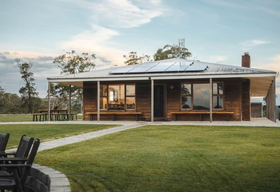 Single-storey wooden house with metal roof and solar panels on a lawn, large windows, benches, outdoor seating, trees in background.