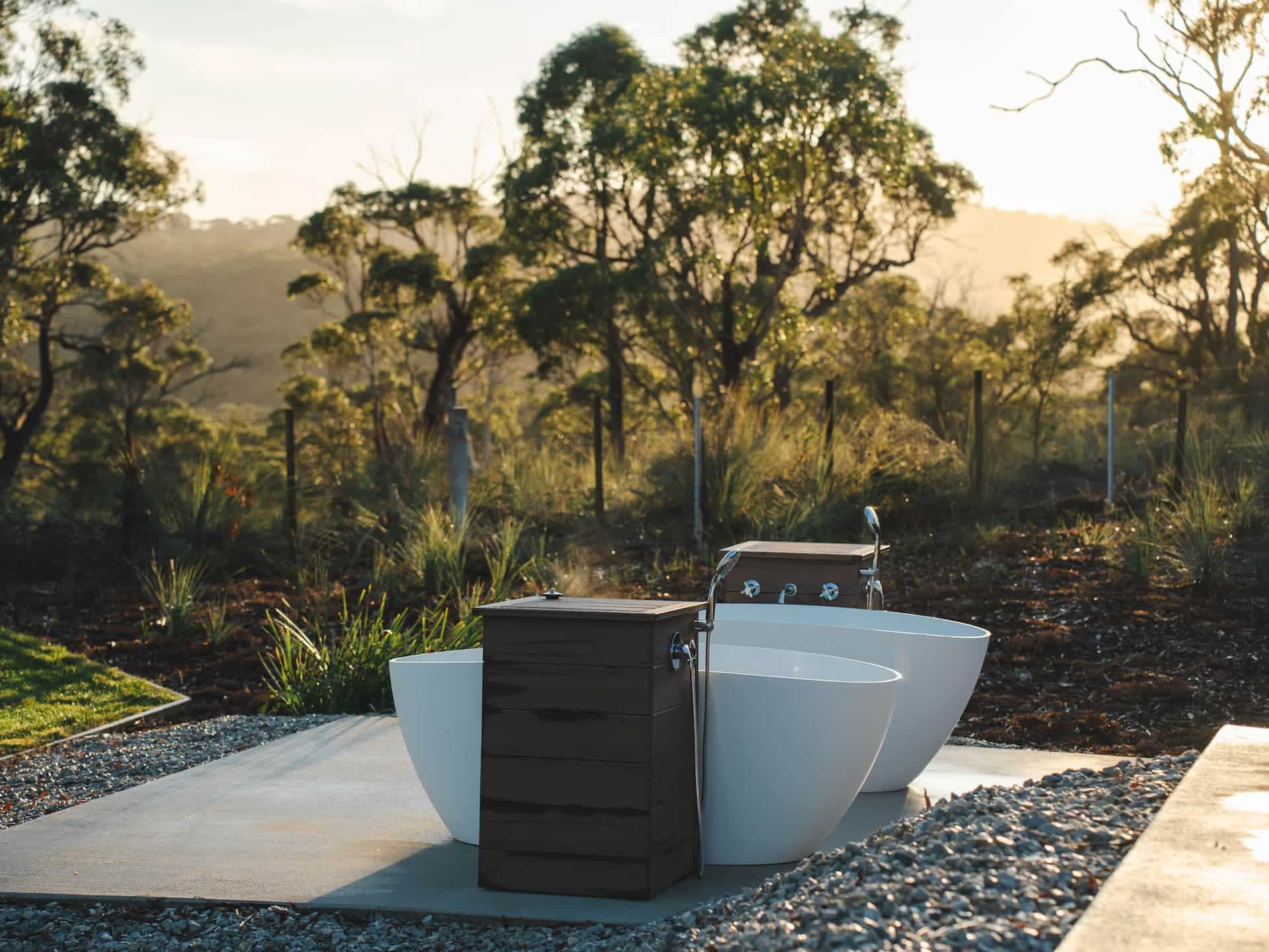 Two white outdoor baths are placed side by side on a concrete slab, surrounded by nature with trees and shrubs in the background.