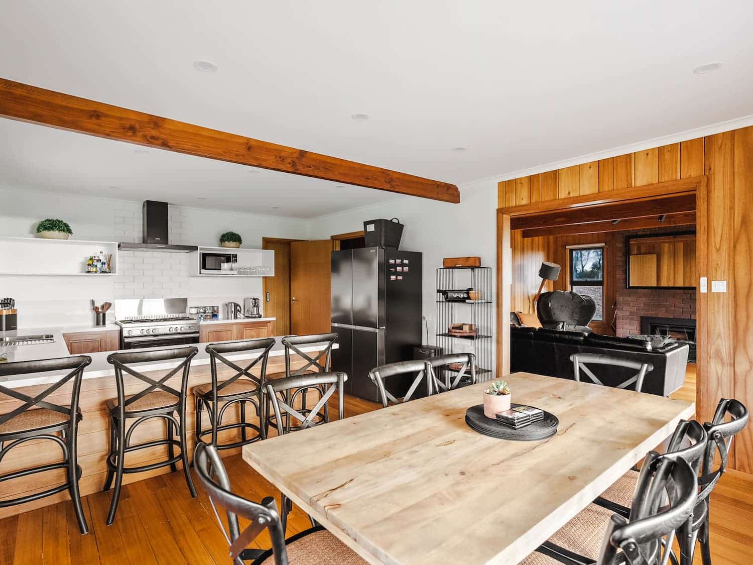Open-plan kitchen and dining area with wooden floors, black chairs, modern appliances, and a view into a cosy living room with wood panelling.