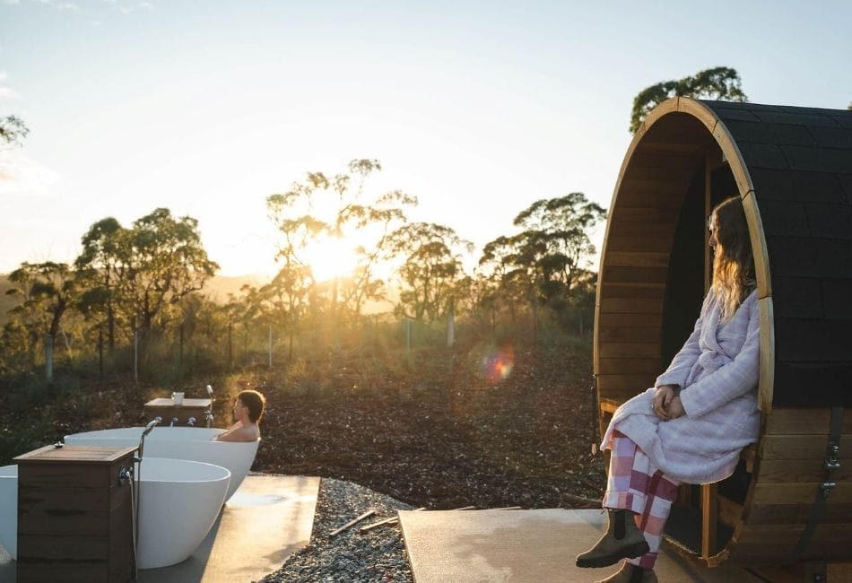 A person in a dressing gown sits on a wooden sauna edge; another person relaxes in an outdoor bath at sunset, trees in the background.