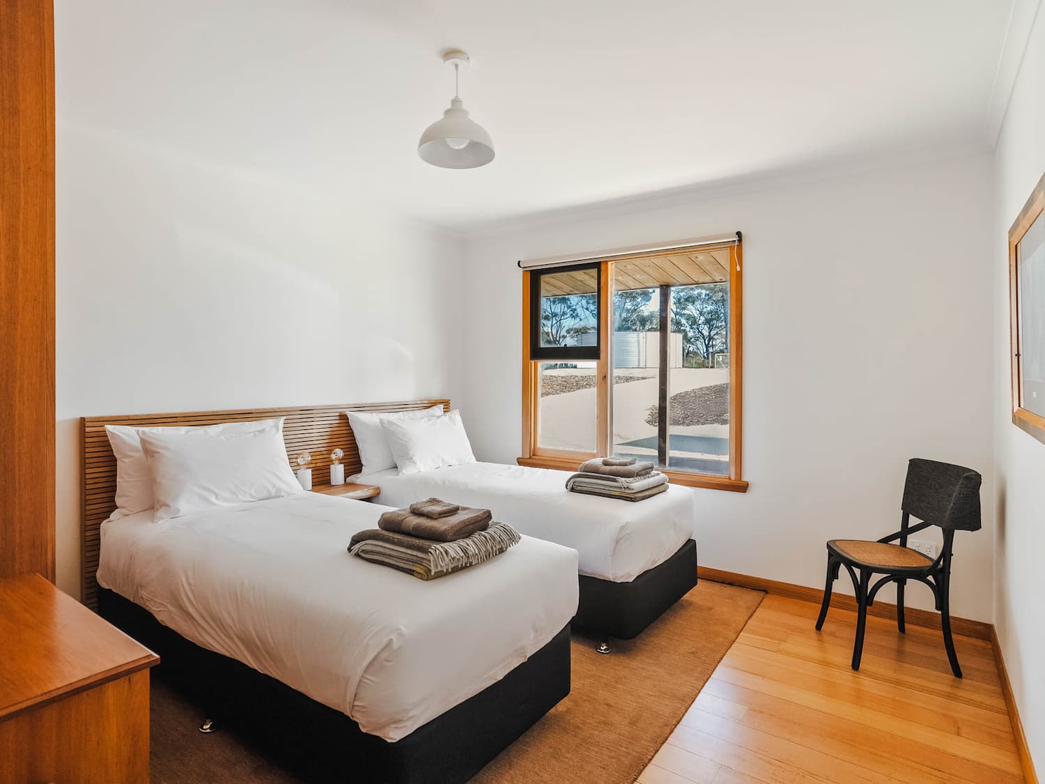 A bright bedroom with two single beds, neatly made with white linen and folded towels, a wooden floor, a window with outdoor view, a black chair, and simple decor.