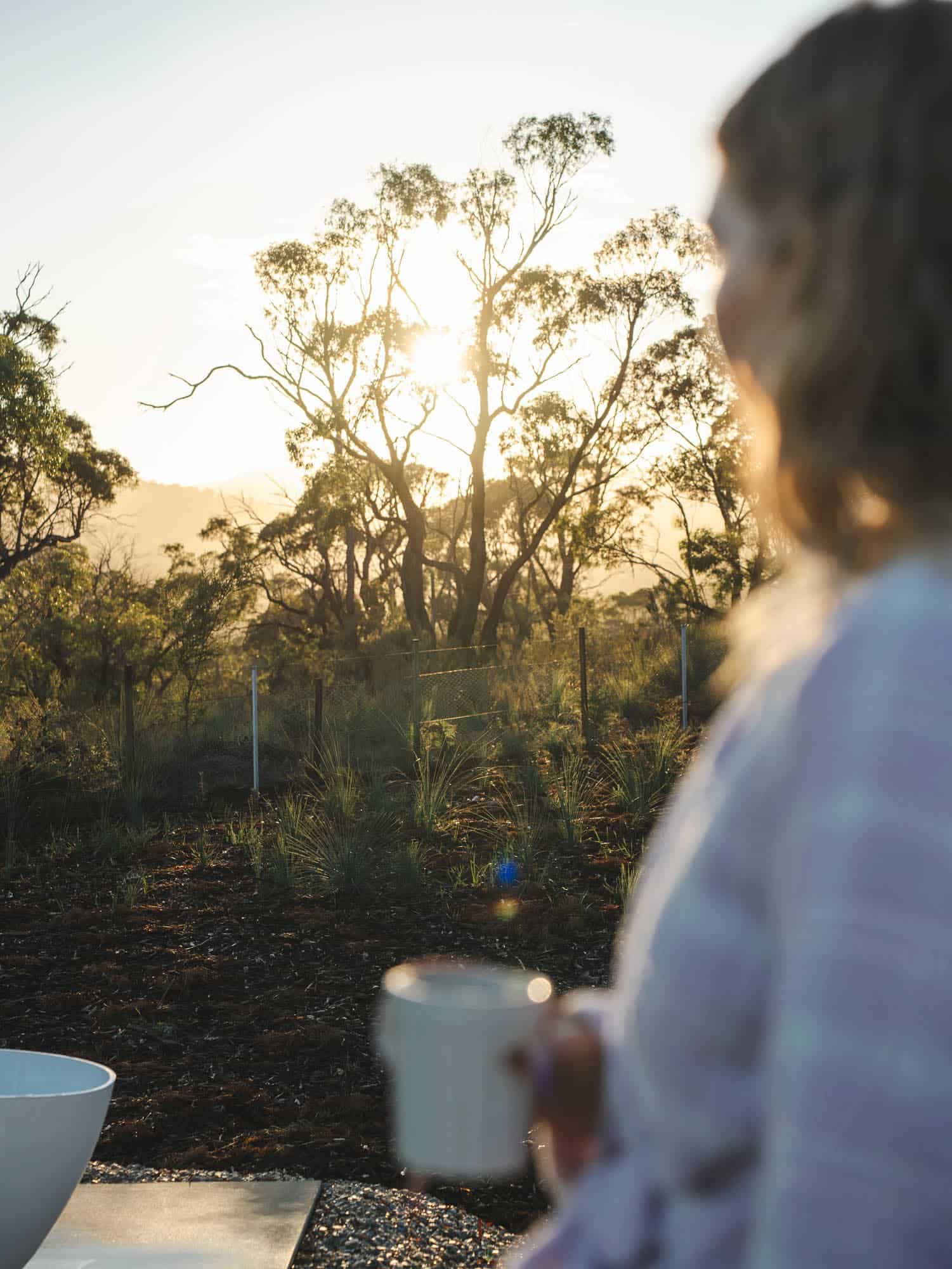 A person holding a mug stands outdoors, out of focus, with sunlight shining through trees in the background.
