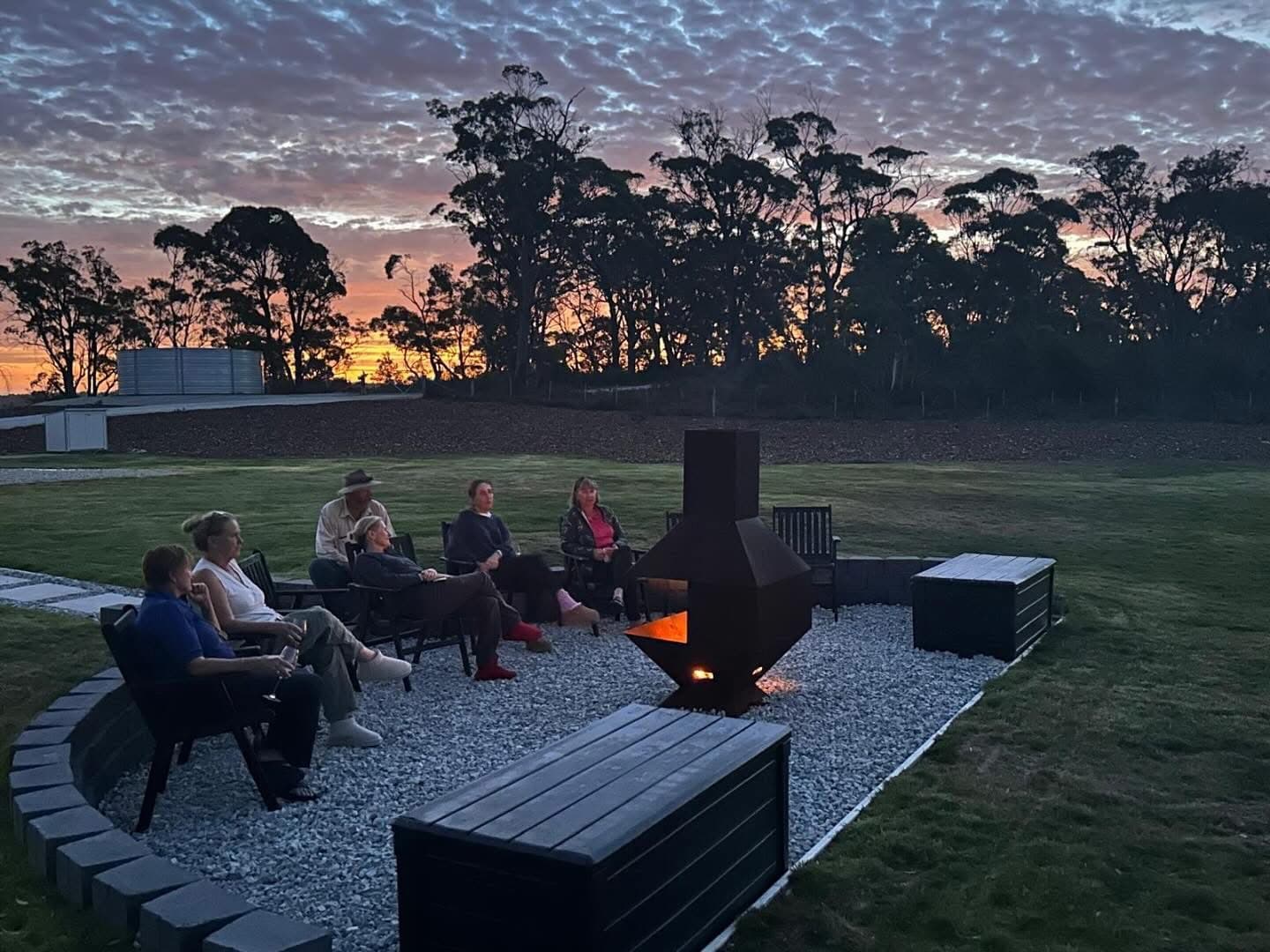 Six people sit around a fire pit on a gravel area in a back garden at dusk, with trees and a dramatic sunset sky in the background.
