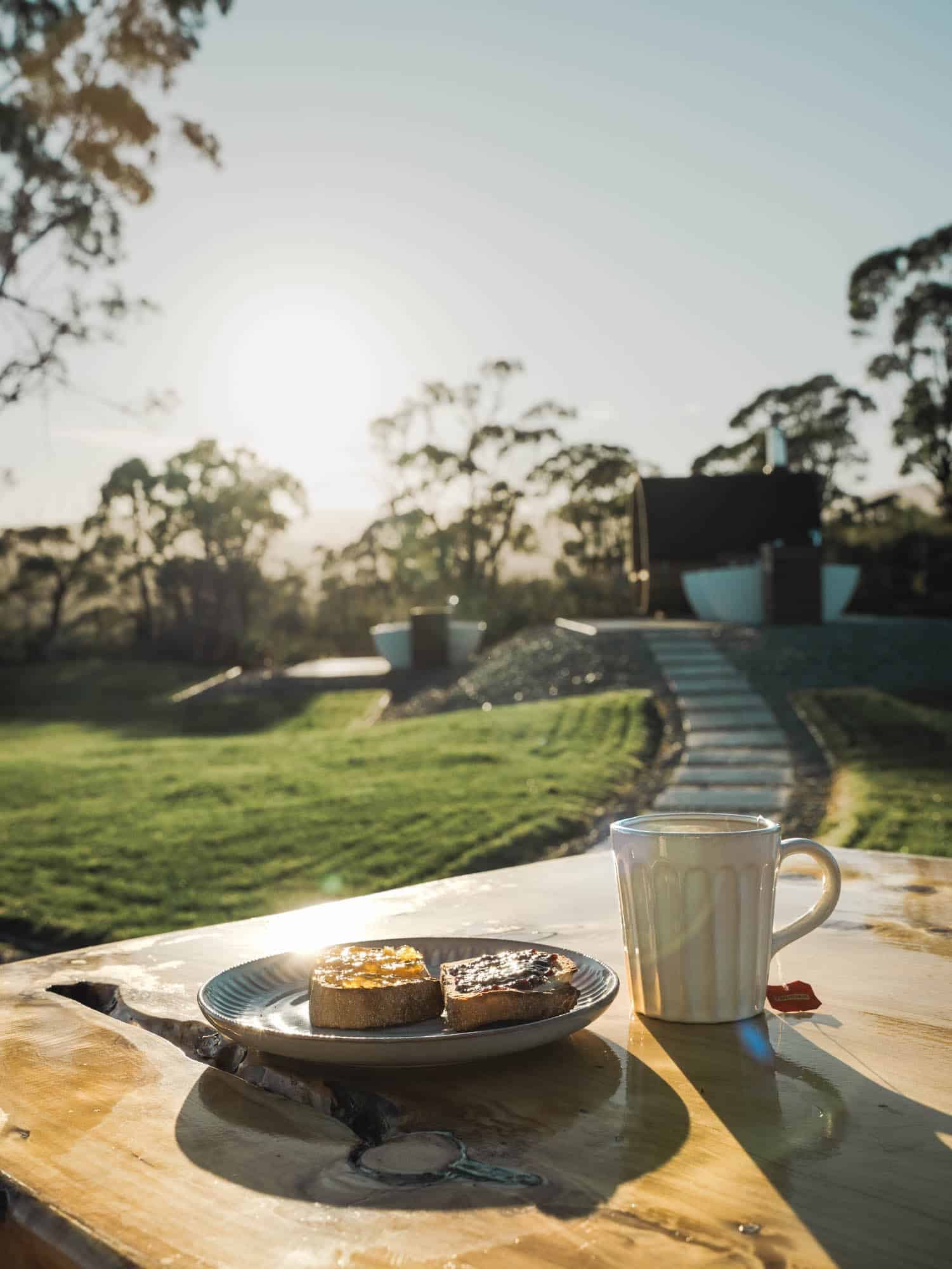 A plate with two pieces of toast and a mug of tea sits on a wooden table outdoors, with a grassy garden and trees in the background at sunrise or sunset.